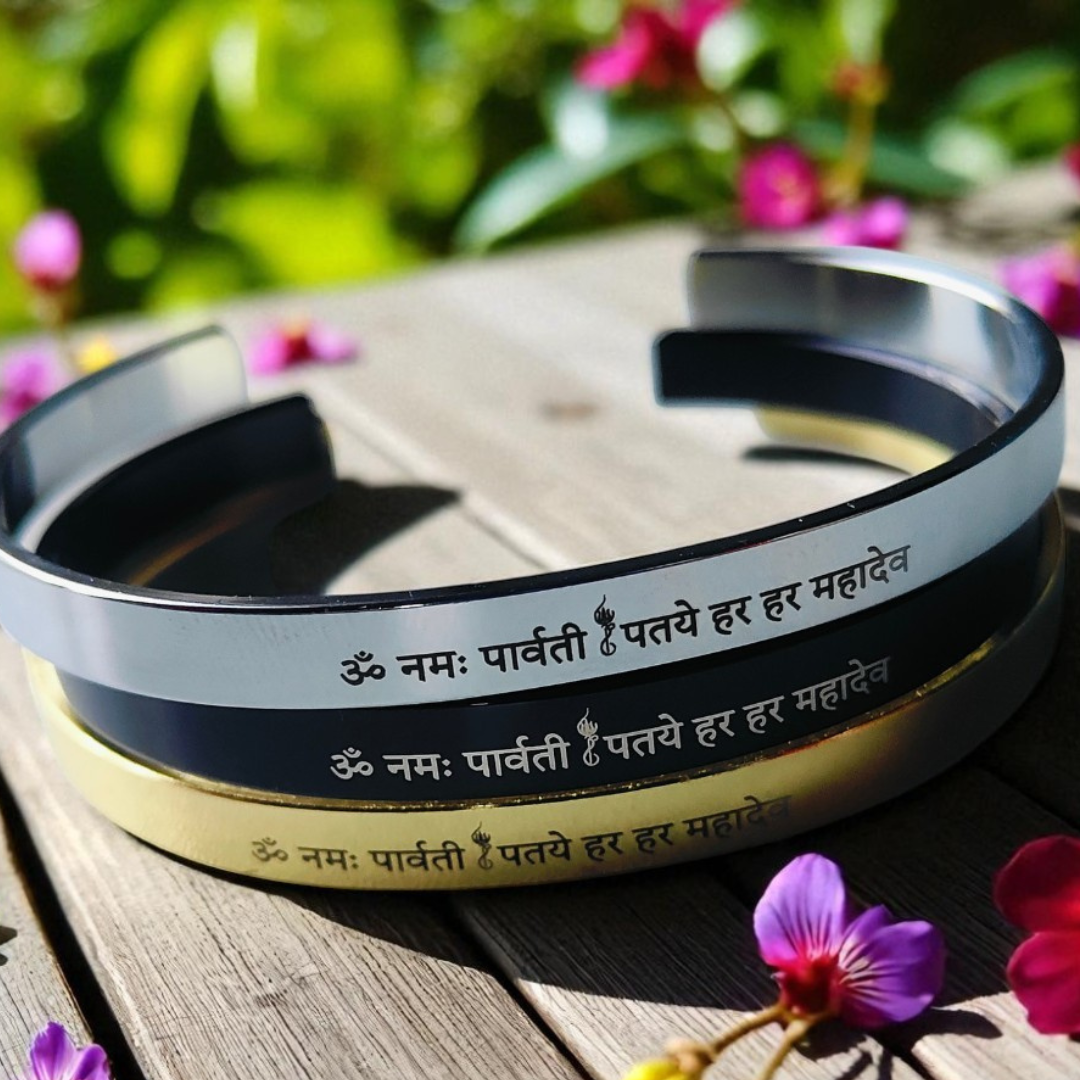 Two metal bracelets with engraved Shiv Mantra on a wooden surface with flowers in the background