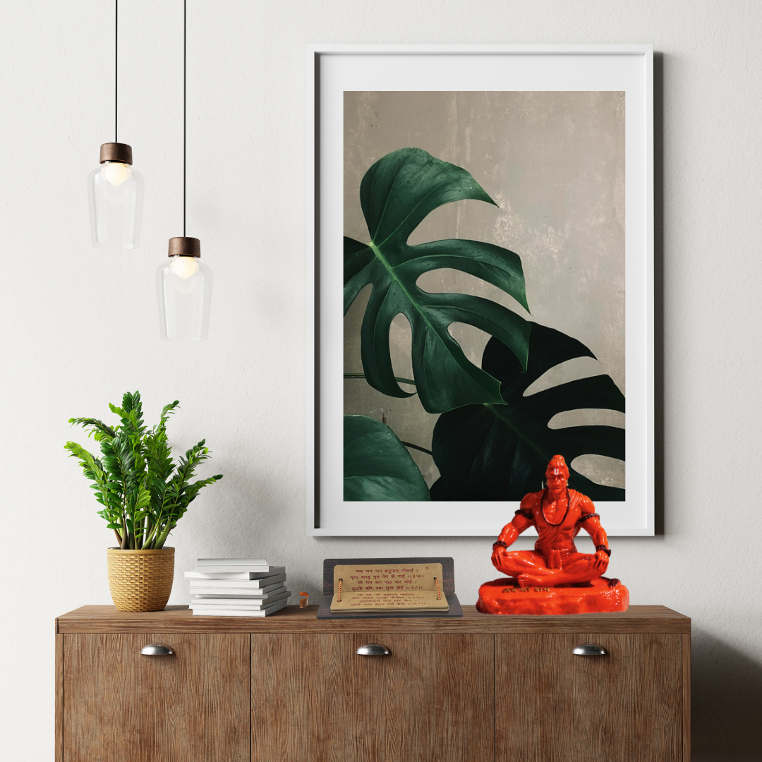 Decorative setup with a framed plant print, orange Hanuman statue, and potted plant on a wooden cabinet.